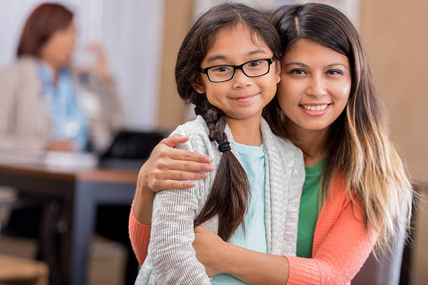 Asian mother and daughter wait in hospital waiting room Mid adult Asian mom embraces elementary age daughter in emergency room waiting area. They have brown hair. The girl is wearing glasses. philippine girl stock pictures, royalty-free photos & images