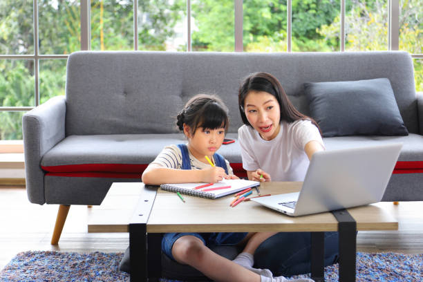 Asian mother working online from home using laptop while watching her daughter working on art class assignment from school for homeschooling and education stock photo