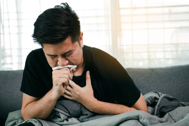 Asian people are sick or ill with bronchitis while coughing by covering their mouth with tissue paper when he sit on the sofa at home. stock photo