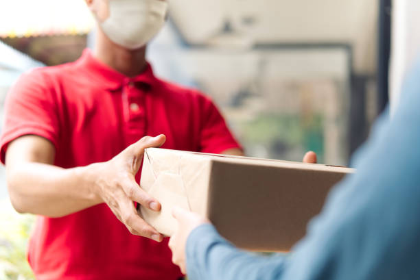 Asian postman, deliveryman wearing mask carry small box deliver to customer in front of door at home. Man wearing mask prevent covid19, corana virus affection outbreak. Social distancing work concept.  package stock pictures, royalty-free photos & images