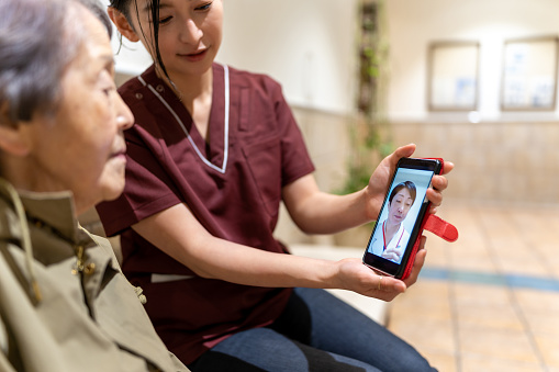 Japanese nursing care staff helps Japanese senior woman watching smart phone. Video call from her doctor. Telemedicine.