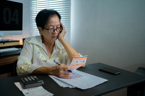 Asian senior woman sit in living room using  calculates monthly expenses, check receipts to pay, do paperwork, busy in part-time retirement job at home, family budget management concept.