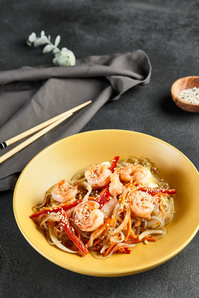 アジアの炒め物エビとライスヌードル。濃いスレートテーブルに木製の箸を入れた黄色い皿にエビ入りのごまライス麺。アジア料理、本格的な食べ物のコンセプト - 日本食 写真 ストックフォトと画像