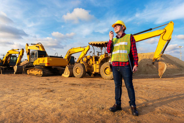 asian technician civil engineer talk with team by walkie talkie hold laptop computer or tablet for transport construction work project with excavator on engineering site background stock photo