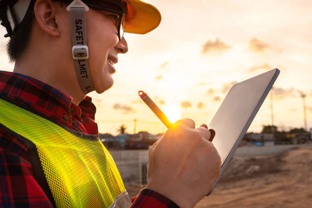 asian technician civil engineer use tablet with smart pen technology on transport site construction to inspect blueprint engineering work online with team at sunset time stock photo