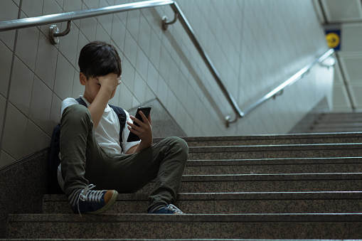 Social issues in internet technology and social media in teenager mental health. Low self esteem young Asian teenager boy sitting alone crying with smartphone, feeling frustration, fear, pain, anxiety, abused as victim of cyberbullying.
