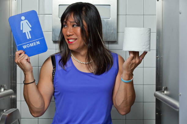 Asian Transgender female happy and smiling holding a women's restroom sign and a roll of toilet paper, Sitting in a restroom stall. stock photo