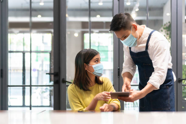 Asian Waiter wearing protective face mask taking order from Female customer on digital tablet in modern cafe stock photo