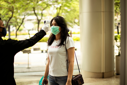 security guard measuring temperature of young woman  with infrared thermometer before entering a office, in Jakarta.