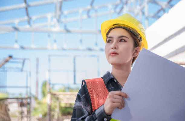 Asian woman civil engineer wear yellow safety hard hat standing and holding blueprint while checking infrastructure of building in construction site. stock photo