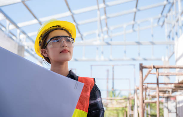 Asian woman civil engineer wears safety yellow helmet with goggles and holding blueprint while check to building in construction site. stock photo