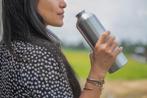 Close-up shot of an Asian woman drinking water from a reusable stainless steel bottle while standing outdoor in the park.