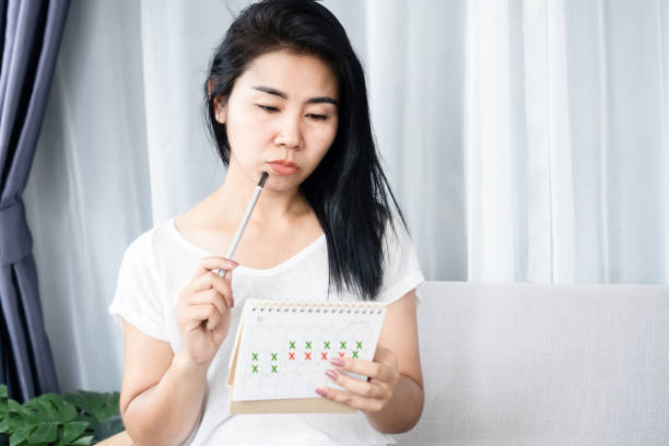 Asian woman having problem with amenorrhea, irregular periods looking at calendar and counting her menstrual cycles stock photo