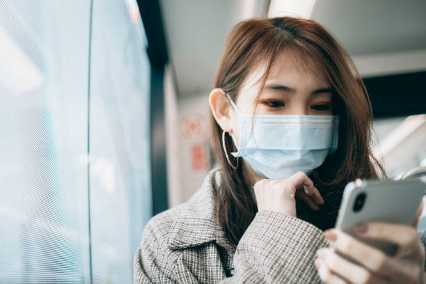亞洲婦女拿著帶手術面罩的智能手機， 在地鐵或火車上通勤時， 保持社會距離圖像檔