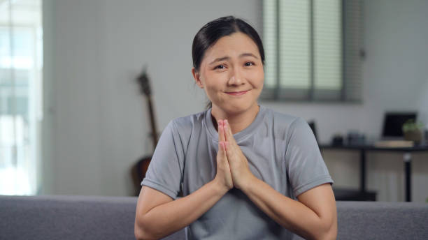 Asian woman holding hands in prayer, looking at camera sitting on sofa at home. stock photo
