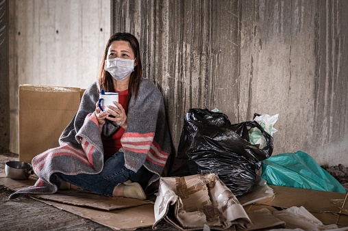 Asian woman homeless wearing hygiene face mask for protective infection and pandemic of coronavirus or covid19. Female beggar wearing sweater and blanket sitting hold bowl for help on walkway street
