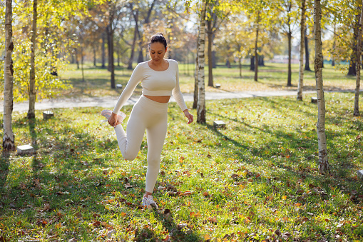 Asian woman in casual sports outfit stretching in the forest, standing on one leg and bending the other backwards holding ankle, leisure outdoor workout on sunny autumn day