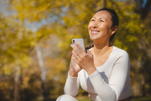Asian woman in casual sports clothes sitting in the park, holding smart phone, smiling and looking up, taking a break from outdoor workout on sunny autumn day
