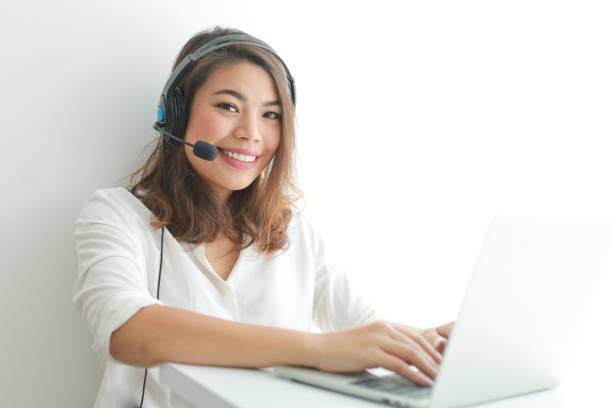 Asian woman on white shirt speak with headphone and using laptop on white background smile and happy face operator concept stock photo