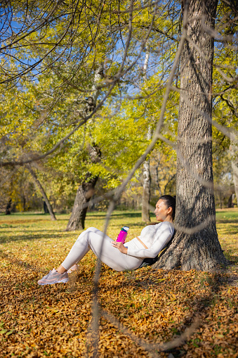 Asian woman in casual sports clothes sitting on dry leaves in the forest, leaning on a tree with water bottle in hands, relaxing after outdoor workout