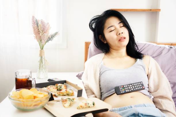 asiatisk kvinna sover i sängen efter att ha ätit pizza, potatischips och läsk med tv-fjärrkontroll på sin feta mage - nap middle age woman bildbanksfoton och bilder