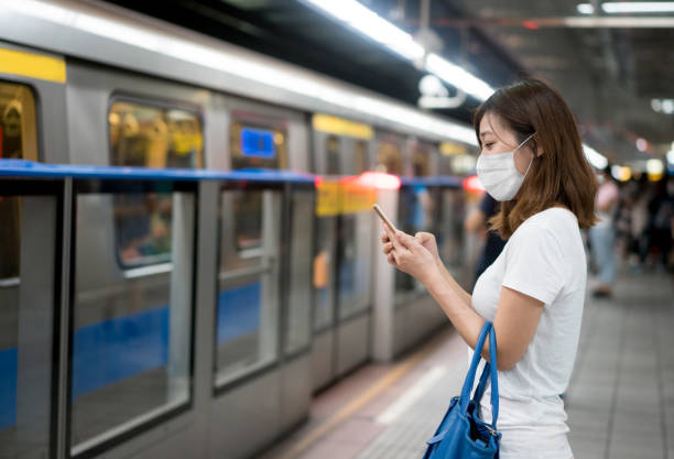 Asian woman texting at the metro station wearing a facemask Asian woman texting at the metro station on her cell phone wearing a facemask to avoid an infectious disease emergency response stock pictures, royalty-free photos & images