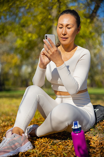 Asian woman in casual sports clothes sitting on dry leaves in the park on sunny day, using mobile phone for social networking