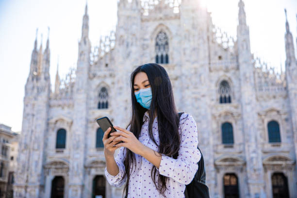 donna asiatica che cammina con una maschera medica usando lo smartphone durante il coronavirus nel duomo di milano - lombardia foto e immagini stock