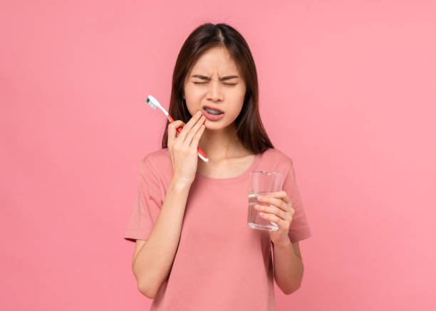 Asian woman wearing braces with brushing teeth with sensitive teeth and holding water glass on pink background, Concept oral hygiene and health care. stock photo