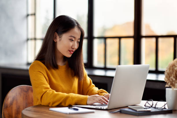 Asian woman working on laptop at home or in cafe. Young lady in bright yellow jumper stock photo