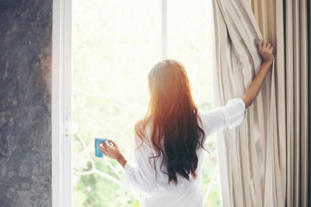 アジアの女性はコーヒーを飲み、彼女のベッドで目を覚まし、日差しの上に新鮮な空気を得るために午前中にカーテンを開けます - beauty and health ストックフォトと画像