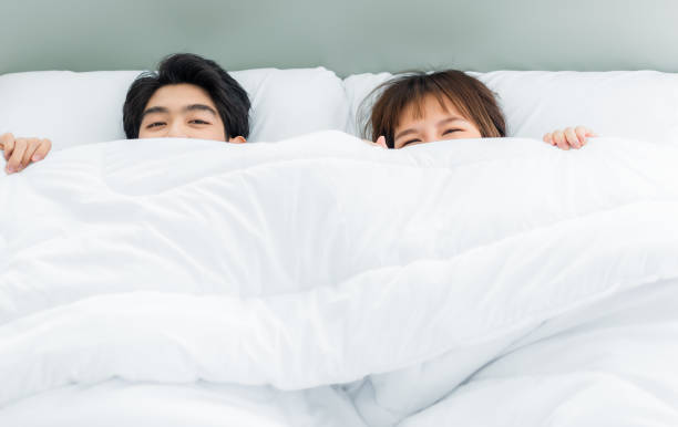 Asian young couple hiding under white blanket on bed stock photo