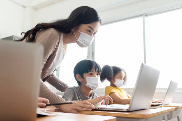Asian Young Teacher with little boy in protective face masks using laptop for  studying in classroom. stock photo