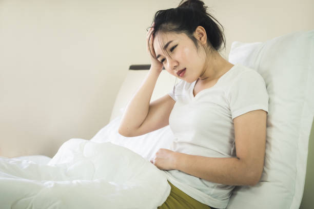 Asian young woman suffering from headache and abdominal pain while sitting on bed at home stock photo