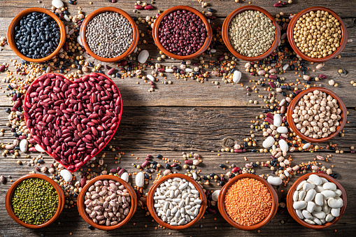 Assorted legumes in pottery bowls with red heart on rustic wooden background frame with mixed legumes including lentils, chickpeas,soybeans and beans