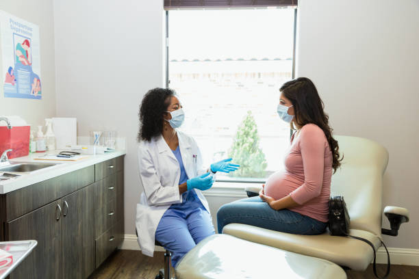 At appointment during COVID-19, doctor gestures and talks to patient At the prenatal appointment during the coronavirus epidemic, the mature adult female doctor with the protective mask gestures and talks to her pregnant patient. obstetrician photos stock pictures, royalty-free photos & images