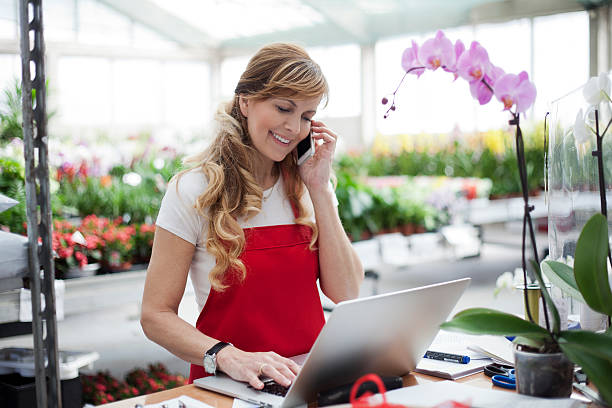 At my garden store Beautiful mature female , smiling works at garden centre retail store.Taking orders over the phone. She working at table and in background is a shop with lots of flowers e commerce assistant stock pictures, royalty-free photos & images