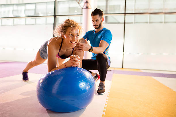 Athletic woman having Pilates training with fitness instructor. Young smiling woman exercising on fitness ball with help of personal trainer in a gym. yoga ball work stock pictures, royalty-free photos & images
