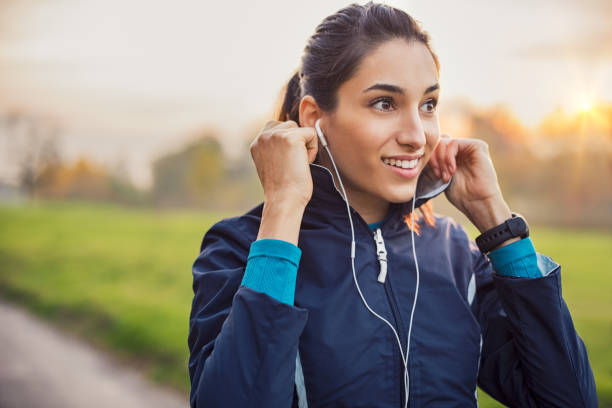 Athletic woman listening music Young athlete adjusting jacket while listening to music at park. Smiling young woman feeling relaxed after a long run during the sunset. Happy sporty woman smiling and looking away during workout. jacket stock pictures, royalty-free photos & images