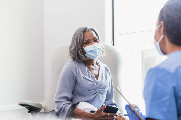 A senior woman holds attentively listens as a female doctor discusses her health. The patient is holding a smartphone. The patient and doctor are wearing protective face masks.