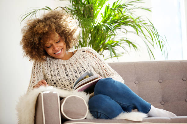 attractive african american woman sitting on sofa reading book - ler imagens e fotografias de stock