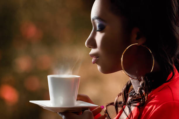 Attractive African woman smelling hot cup of coffee. Close up side view of attractive african woman smelling scent of hot beverage.Low key portrait of woman with directional backlit ambient light holding cup with coffee. scented stock pictures, royalty-free photos & images