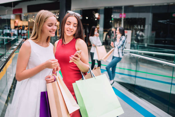 Attractive and beautiful girls are standing together with bags. Brunette girl is holding phone and looking at it together with her friend. Blonde girl is smiling. Their friends are standing behind. stock photo