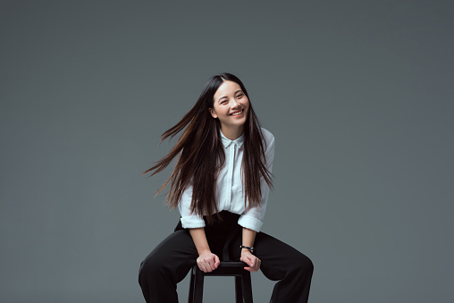 attractive asian girl sitting on chair and smiling at camera isolated on grey
