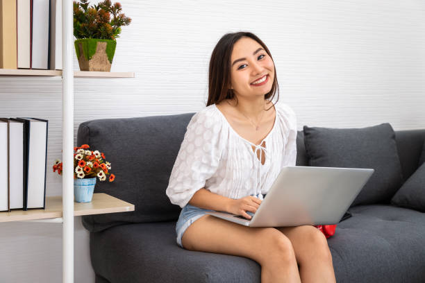 Attractive Asian woman sitting on sofa couch in her living room using laptop to work from home, looking at camera stock photo