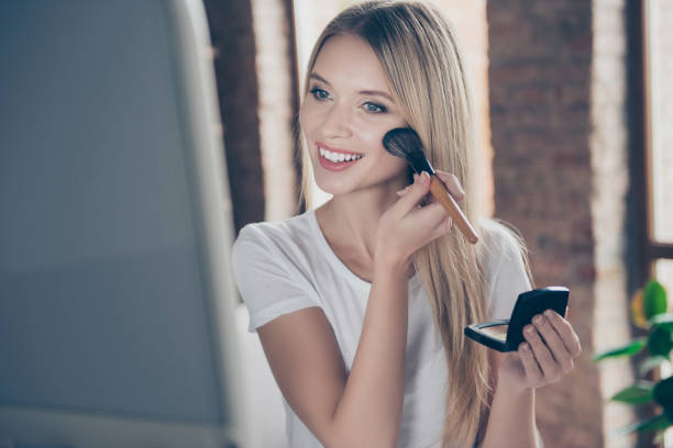 Attractive beautiful charming joyful glad woman wearing white t-shirt is applying blusher on her cheekbones in front of mirror at home Attractive beautiful charming joyful glad woman wearing white t-shirt is applying blusher on her cheekbones in front of mirror at home blusher make up stock pictures, royalty-free photos & images