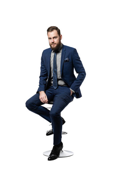Attractive Caucasian businessman wearing an elegant blue suit and sitting on a stool Confident, attractive and
bearded CEO looking elegant in his custom made blue suit and
tie, with one hand in his pocket and sitting on a stool, isolated on
white background. stool stock pictures, royalty-free photos & images