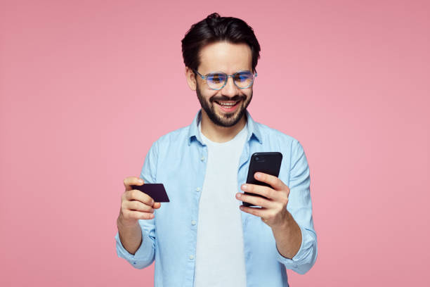 Attractive handsome man holding plastic credit card and mobile phone isolated over pink background. Closeup portrait of young bearded man holding plastic credit card and mobile phone isolated over pink background. Stylish male making shopping on smartphone credit card photos stock pictures, royalty-free photos & images