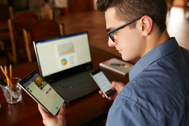 attraktiva mannen i glasögon arbetar med flera elektroniska internet enheter. freelancer affärsman har bärbar dator och smartphone i händerna och laptop på bord med diagram på skärmen. multitasking tema - utrustning bildbanksfoton och bilder