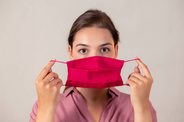 Attractive medical worker about to put on an earloop face mask Young girl looking at the camera and preparing to put on a mask. Woman on a light background putting on a medical antiviral protection. getting dressed stock pictures, royalty-free photos & images